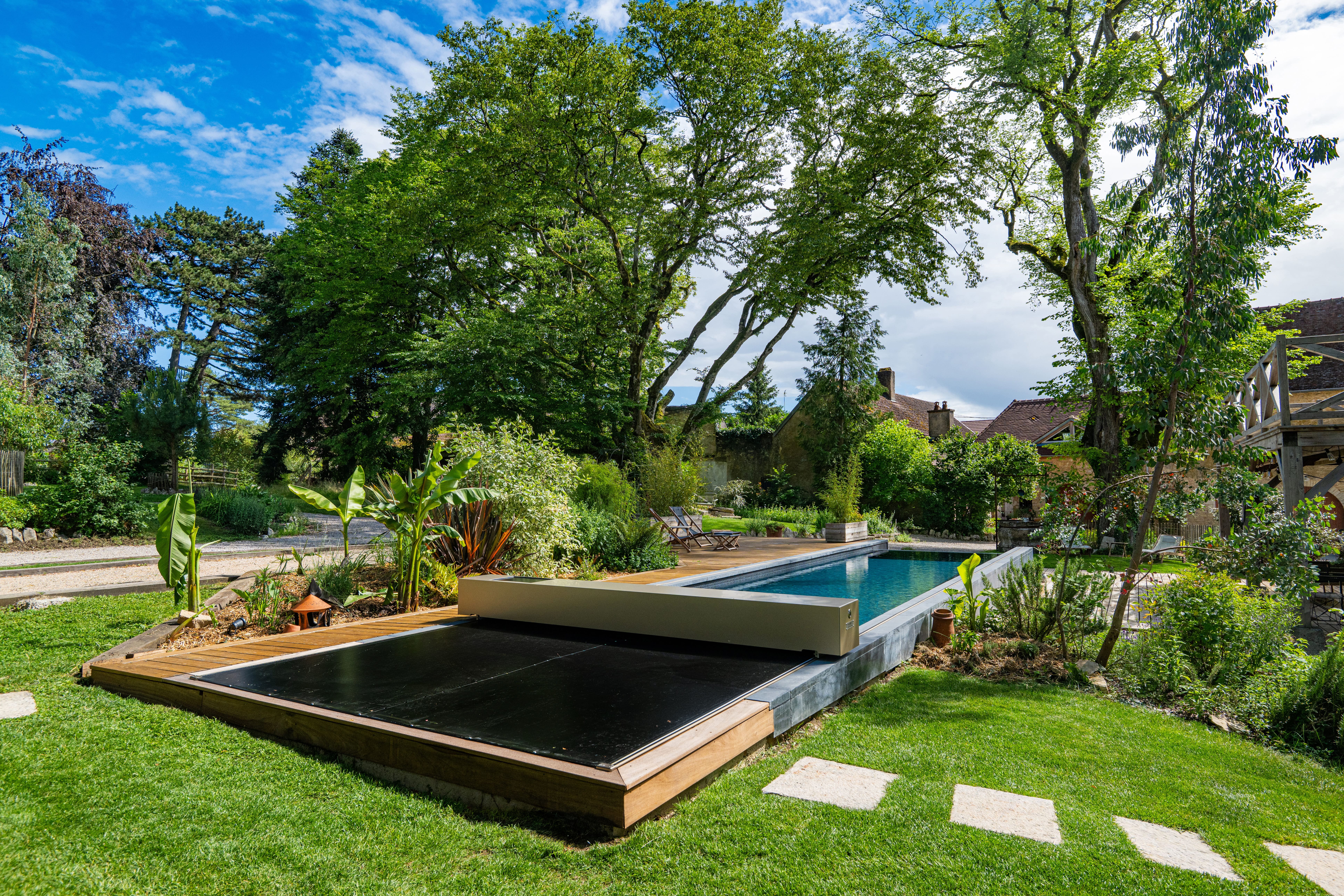 Piscine extérieure partiellement recouverte d’une membrane Coverseal noire, intégrée à un jardin verdoyant avec terrasse en bois.